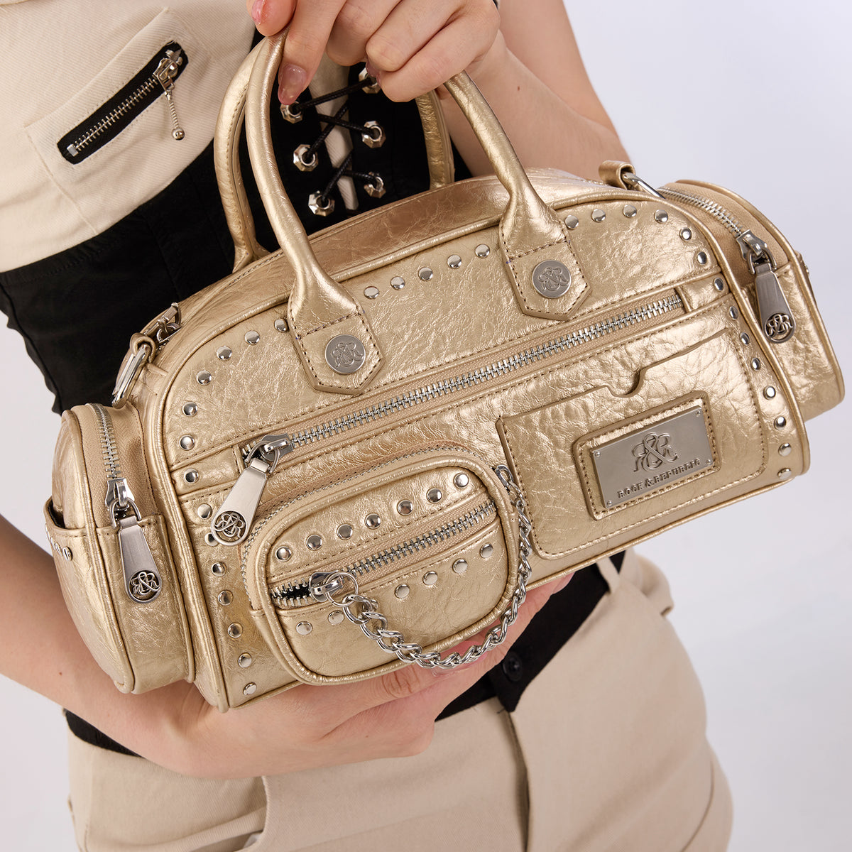 Rock & Republic Studded Mini Boston Bag in Gold PU held in hands showing studded pockets and structured boston profile.