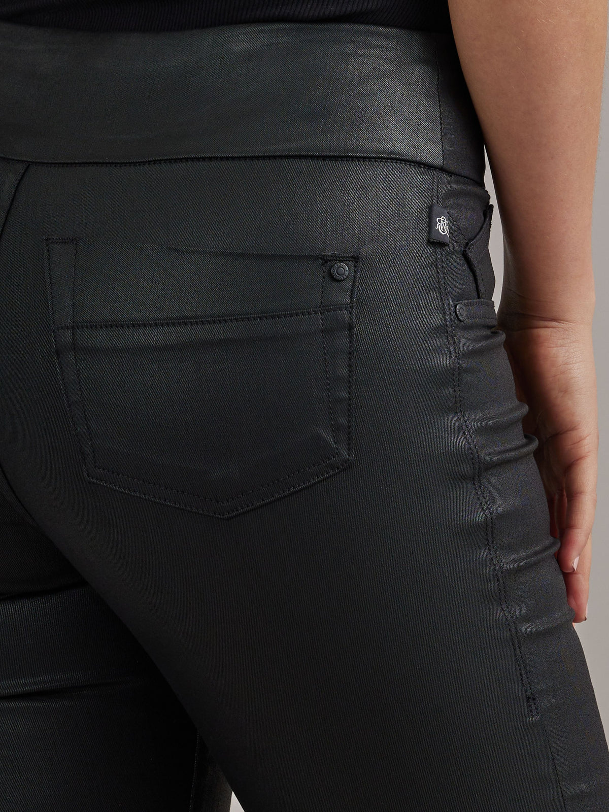 Side waist detail of Rock & Republic Women's Fever Legging in Blacklist showing coated denim, rivet, and RR logo tab.