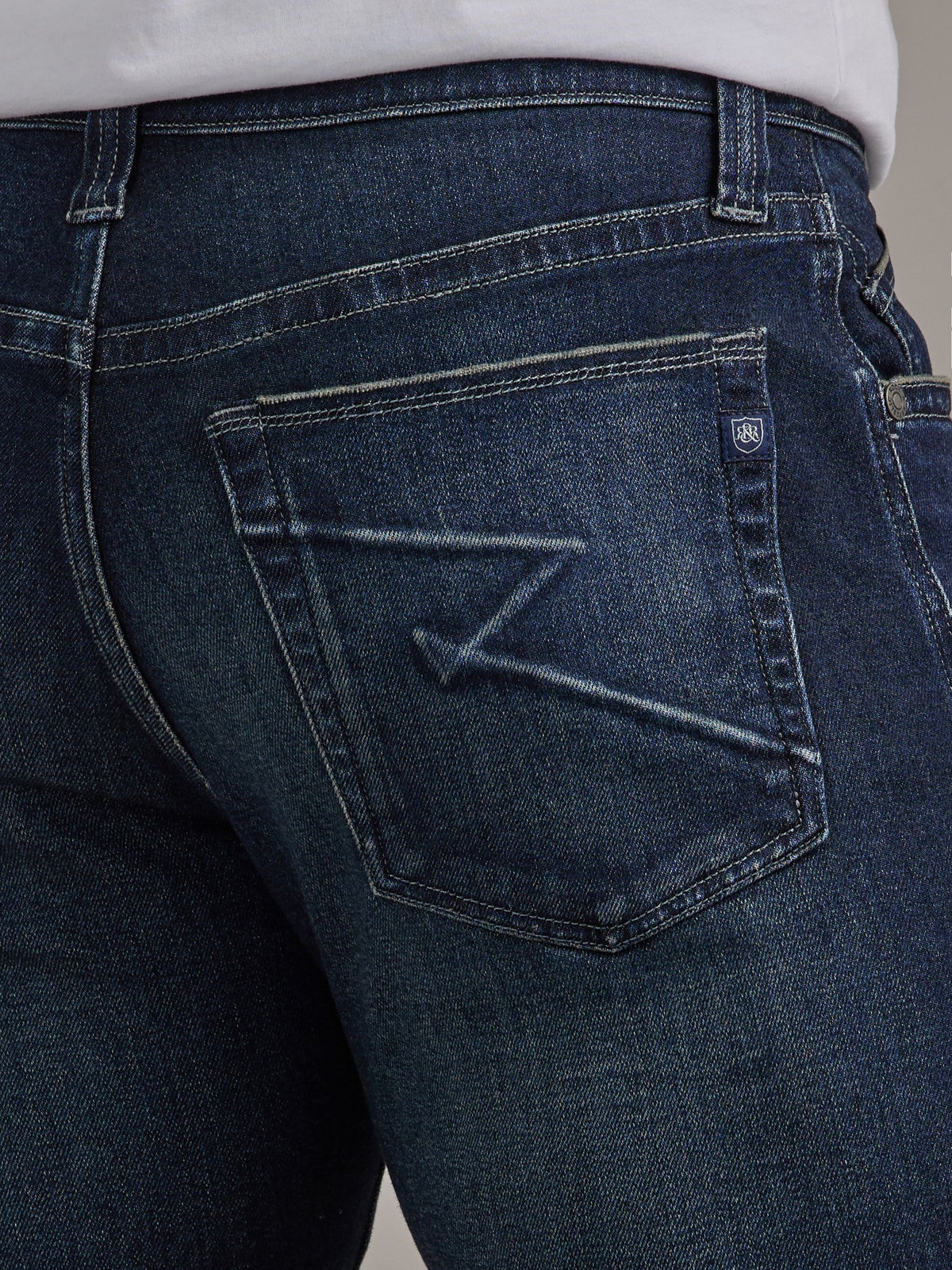 Detailed back pocket view of Rock & Republic Colburg jeans in Salute showing geometric creased design and crest logo tab.