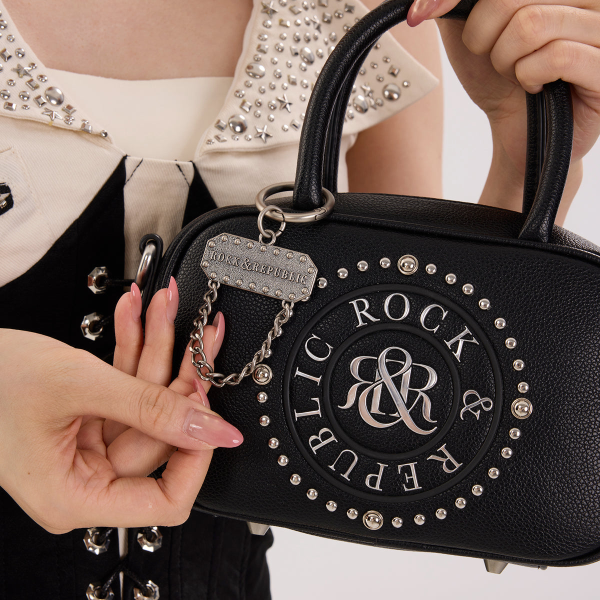 Rock & Republic Studded Logo Medallion Satchel Bag in Black PU detail view showing medallion on satchel profile.