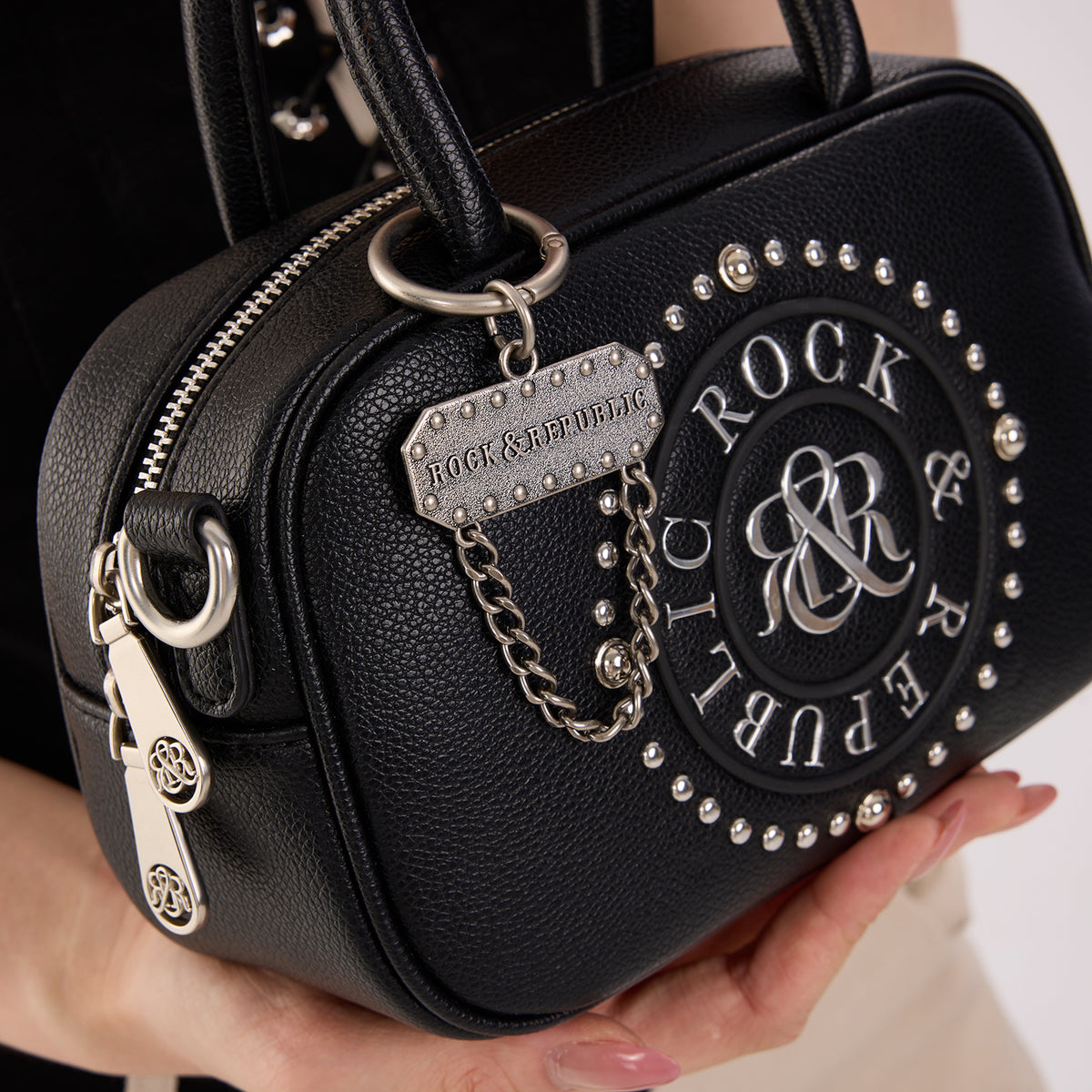 Rock & Republic Studded Logo Medallion Satchel Bag in Black PU close-up highlighting charm on satchel silhouette.